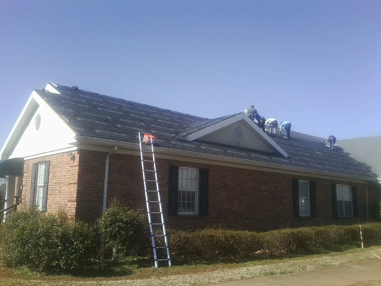 Crew tearing shingles off a brick ranch roof with a ladder set — Emergency Roof Repair work by our crew in Goose Lake