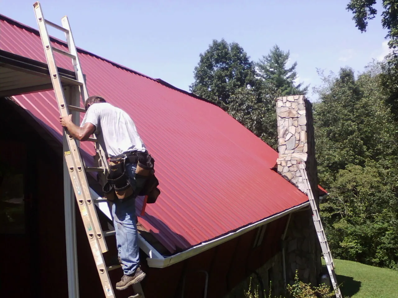 Roofer on a ladder beside a red metal roof and stone chimney — Soffit & Fascia Repair by Roofing Brothers in Goose Lake